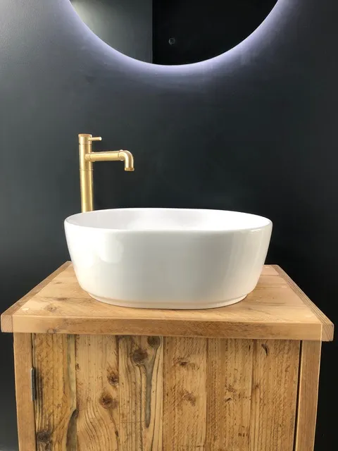Modern Round Ceramic Basin with Gold Tap on Rustic Wooden Vanity | AC Plumbers Ltd. White round ceramic countertop basin with a gold mixer tap, mounted on a rustic wooden vanity unit in a contemporary bathroom.